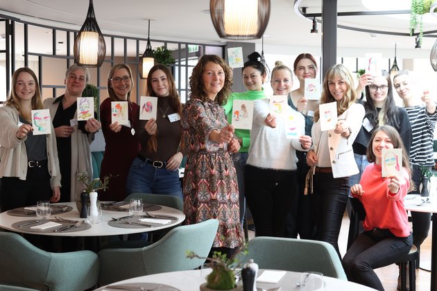Gruppe von 13 Frauen in einem modernen Restaurant, die bunte gezeichnete Karten in die Kamera halten und lächeln.