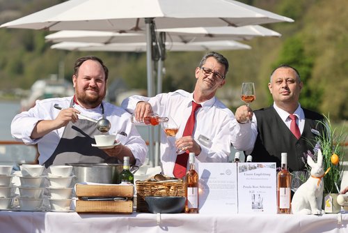 Drei Männer in Arbeitskleidung an einem Tisch mit Suppenschüsseln, Weinflaschen und einem weißen Hasen-Dekor, einer schöpft Suppe, ein anderer gießt Roséwein in ein Glas.