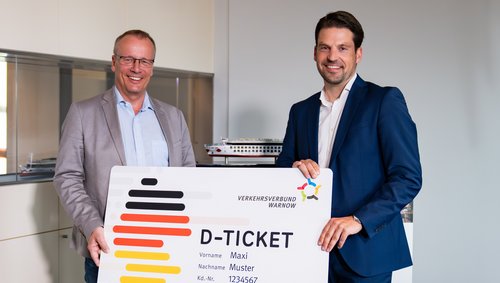 Jörg Eichler und ein weiterer Mann halten ein großes Modell eines D-Tickets des Verkehrsverbund Warnow mit Namen Maxi Muster und Kundennummer 1234567.