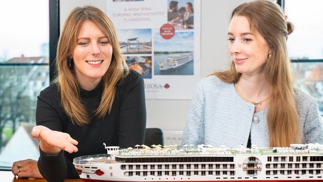Zwei Frauen sitzen an einem Tisch und betrachten ein detailliertes Modell eines Flusskreuzfahrtschiffs in einem hellen Raum mit Fenster und Poster im Hintergrund.
