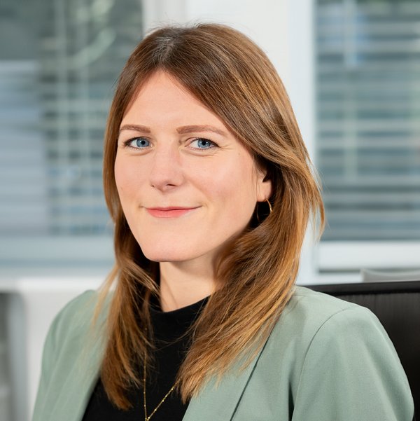 Nadine Richter mit schulterlangem braunem Haar, trägt grünen Blazer und schwarze Bluse, lächelt leicht vor Bürofenstern.