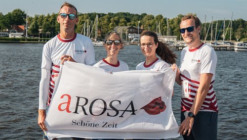 Vier A-ROSA Mitarbeiter, darunter Stefan Sprunk, halten eine weiße Fahne mit dem Schriftzug A-ROSA Schöne Zeit vor einem See mit Booten im Hintergrund.