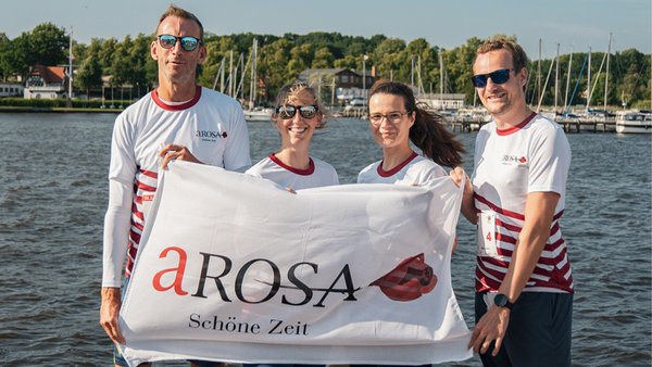 Vier A-ROSA Mitarbeiter, darunter Stefan Sprunk, halten eine weiße Fahne mit dem Schriftzug A-ROSA Schöne Zeit vor einem See mit Booten im Hintergrund.