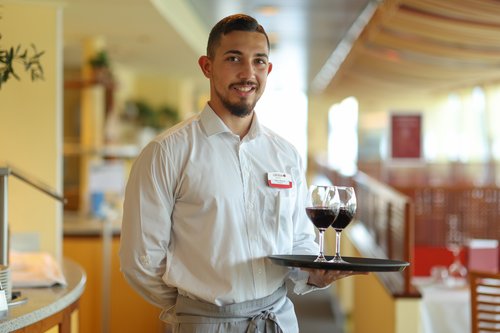 Junger Kellner mit weißem Hemd und Namensschild hält Tablett mit zwei Gläsern Rotwein in einem Restaurant