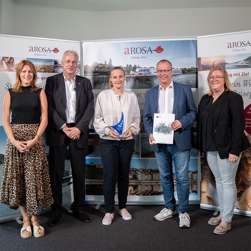 Fünf Personen, darunter Jörg Eichler, stehen vor Bannern mit dem Logo A-ROSA Schöne Zeit. Zwei halten eine Auszeichnung und eine Urkunde für Top-Ausbildungsbetrieb in der Hand.