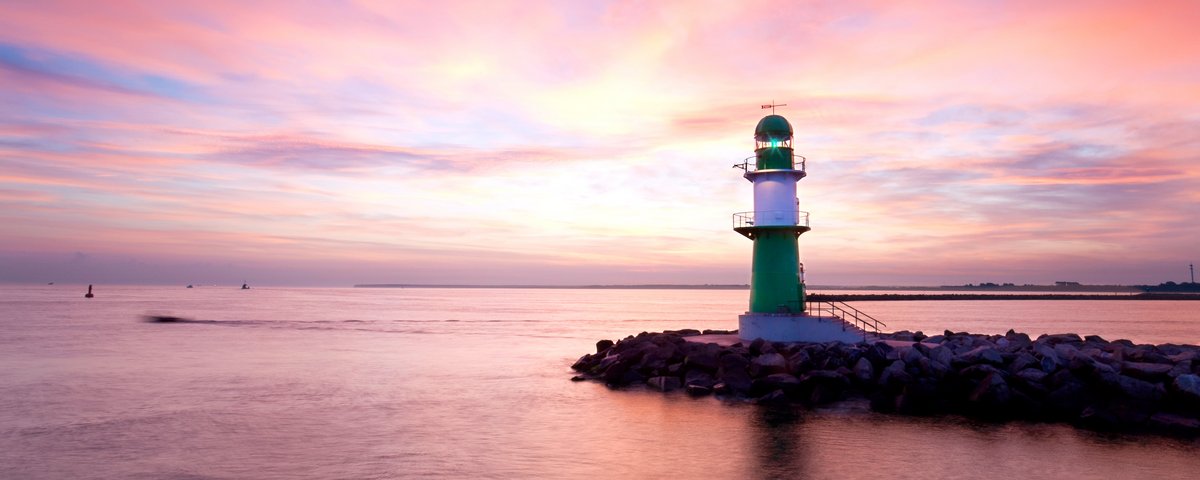 Grüner und weißer Leuchtturm auf Felsen im Wasser bei Sonnenuntergang in Warnemünde mit rosa und orangefarbenem Himmel