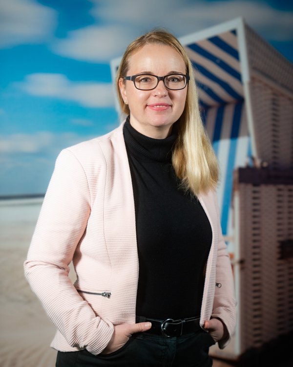Anne Riesner-Koch mit Brille, blondem Haar, schwarzem Rollkragenpullover und hellem Blazer vor Strandkorb und blauem Himmel.