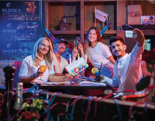 Vier junge Erwachsene sitzen an einem Tisch, stoßen mit Getränken an und machen ein Selfie, im Hintergrund Werbetafeln mit den Worten Klock 8, Disco und Sportclub. Einer hält eine große LOKA Karte in der Hand.