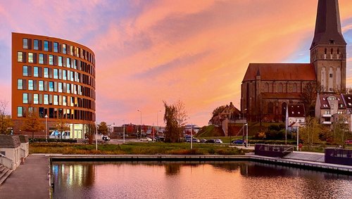 Blick auf das Bürogebäude von A-ROSA in Rostock neben einer Kirche mit hohem Turm bei Sonnenuntergang, Wasser im Vordergrund reflektiert den orangefarbenen Himmel.