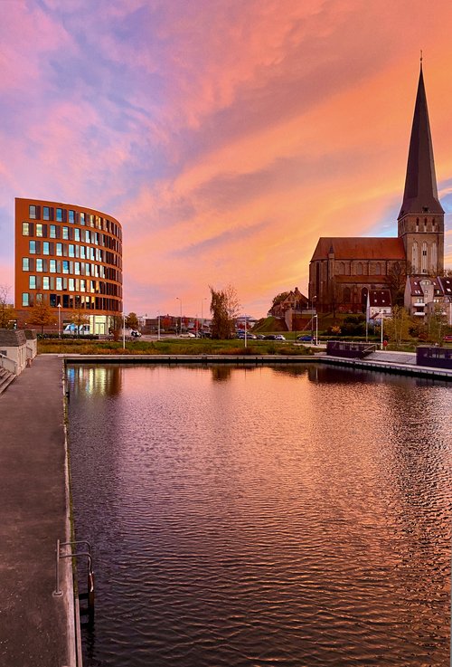 Blick auf das Bürogebäude von A-ROSA in Rostock neben einer Kirche mit hohem Turm bei Sonnenuntergang, Wasser im Vordergrund reflektiert den orangefarbenen Himmel.