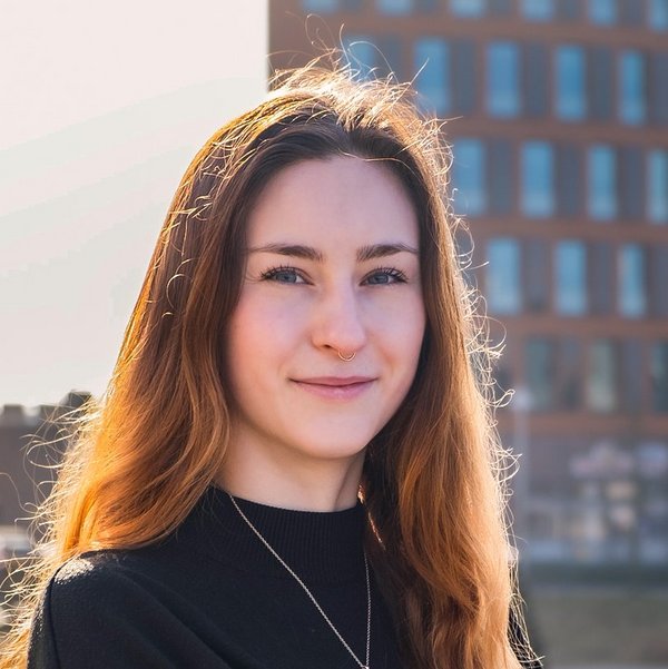 Junge Frau mit langen roten Haaren und schwarzem Pullover lehnt an einem Geländer vor einem modernen, runden Bürogebäude am Wasser.