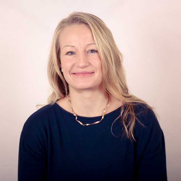 Frau mit langen blonden Haaren, blauem Oberteil und goldener Kette, lächelt in die Kamera vor hellem Hintergrund.