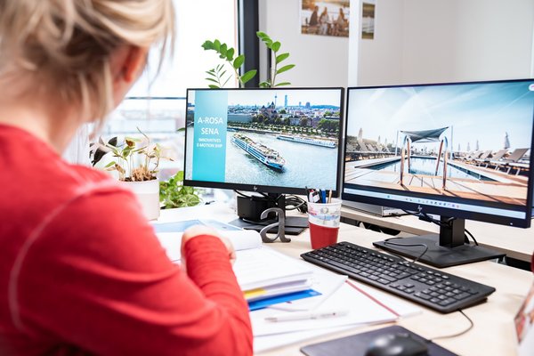 Frau im roten Pullover arbeitet an einem Schreibtisch mit zwei Monitoren, auf einem Bildschirm ist ein Flusskreuzfahrtschiff mit dem Text A-ROSA SENA Innovatives E-Motion Ship zu sehen.