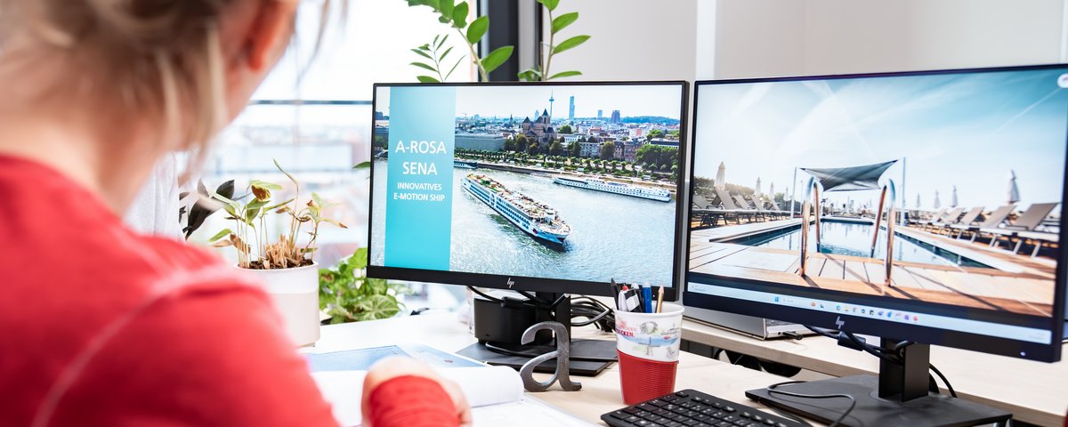 Frau im roten Pullover arbeitet an einem Schreibtisch mit zwei Monitoren, auf einem Bildschirm ist ein Flusskreuzfahrtschiff mit dem Text A-ROSA SENA Innovatives E-Motion Ship zu sehen.