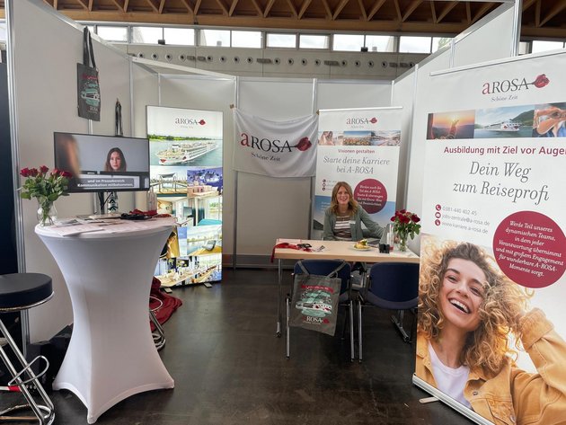 Messestand von A-ROSA mit Bannern zur Karriere und Ausbildung im Reisebereich, Frau sitzt am Tisch, Monitor zeigt Video mit Untertiteln, Rosen auf Tischen