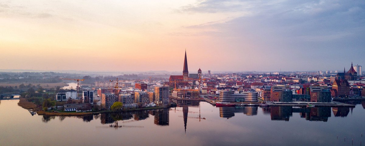 Panorama von Rostock bei Sonnenaufgang mit Stadtansicht, Hafen und markanter Kirchturmspitze.