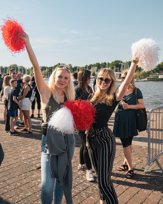 Zwei junge Frauen mit roten und weißen Pompons feiern am Wasser bei sonnigem Wetter, im Hintergrund weitere Menschen und Boote.