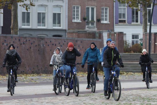 Sieben Personen fahren bei kühlem Wetter auf Fahrrädern in einer Stadt mit historischen Gebäuden im Hintergrund.