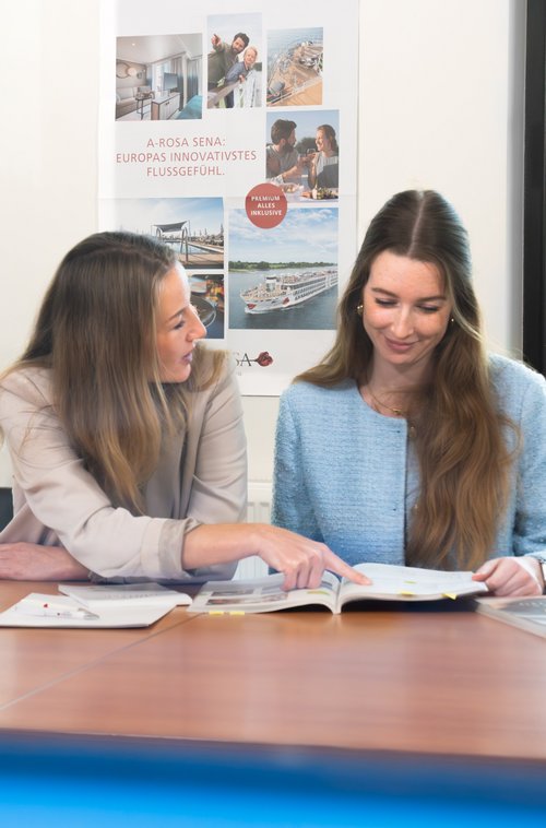 Zwei Frauen sitzen an einem Tisch vor großen Fenstern, eine zeigt auf ein geöffnetes Buch, im Hintergrund ein Poster mit Flusskreuzfahrtschiff und Stadtblick.