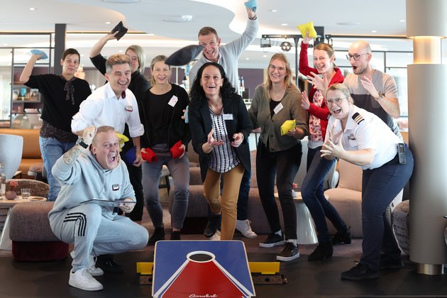 Elf Personen spielen drinnen Cornhole, werfen bunte Säcke auf ein Spielbrett und zeigen Freude und Begeisterung.