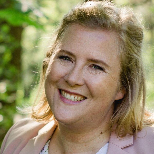 Lächende Frau mit blonden Haaren, trägt hellrosa Blazer, vor grünem, unscharfem Hintergrund.