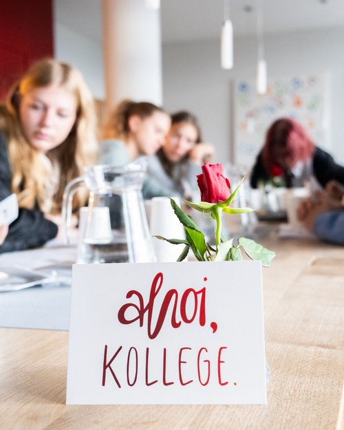 Karte mit roter Schrift Ahoi, Kollege. und rote Rose auf einem Tisch, im Hintergrund unscharfe Personen bei einem Treffen.