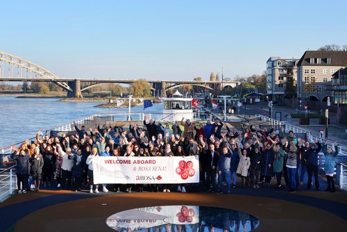 Große Gruppe von Menschen auf einem Flusskreuzfahrtschiff hält ein Banner mit der Aufschrift WELCOME ABOARD A-ROSA SENA! und winkt, im Hintergrund Brücke und Flussufer