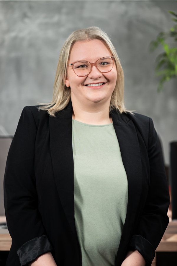 Elisa Gunert lächelnd mit blonden Haaren, Brille, grünem Shirt und schwarzem Blazer vor grauem Hintergrund