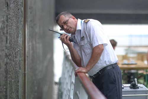 Mann in weißem Hemd mit Schulterklappen spricht in Walkie-Talkie und lehnt sich an ein Geländer