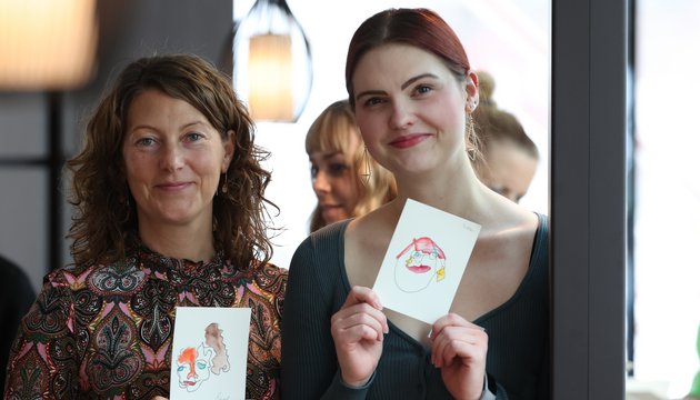 Zwei Frauen lächeln und halten jeweils eine kleine Zeichnung mit abstrakten Gesichtern in der Hand.