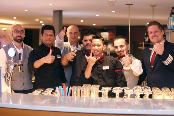 Sieben männliche Barkeeper und Kellner in schwarzer und grauer Arbeitskleidung mit roten Fliegen und Krawatten stehen hinter einer Bar mit Gläsern und bunten Trinkhalmen.