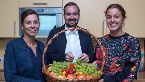 Drei lächelnde Personen in einer Küche halten gemeinsam einen großen Korb mit Trauben, Erdbeeren und anderen Früchten