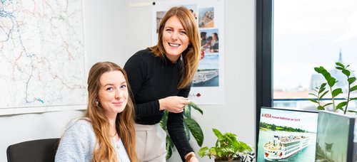 Zwei Frauen im Büro, eine sitzt und die andere steht, beide lächeln. Auf dem Tisch liegen Broschüren und der Monitor zeigt ein Flusskreuzfahrtschiff von A-ROSA.