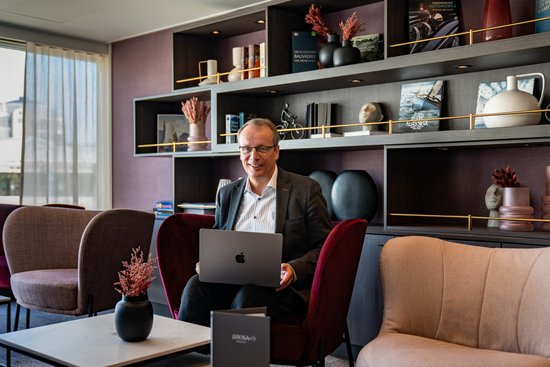 Jörg Eichler sitzt in einem modernen Büro auf einem roten Sessel mit einem Apple Laptop auf dem Schoß, umgeben von bequemen Sesseln und dekorativen Regalen mit Büchern und Vasen.