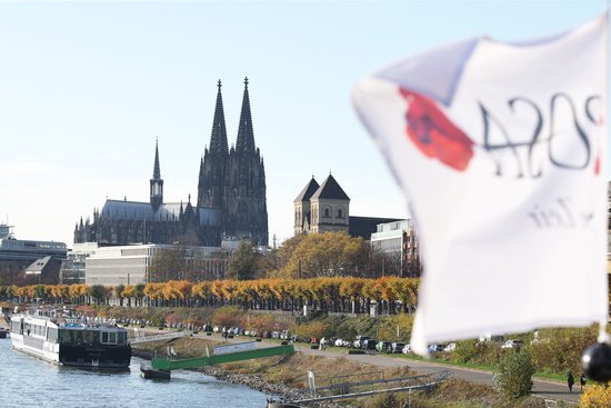 Kölner Dom mit zwei hohen Türmen und umliegenden Gebäuden am Rhein, im Vordergrund ein weißes, unscharfes Fahnenstück