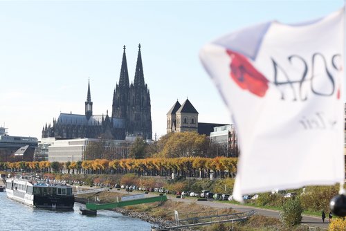 Kölner Dom mit zwei hohen Türmen und umliegenden Gebäuden am Rhein, im Vordergrund ein weißes, unscharfes Fahnenstück